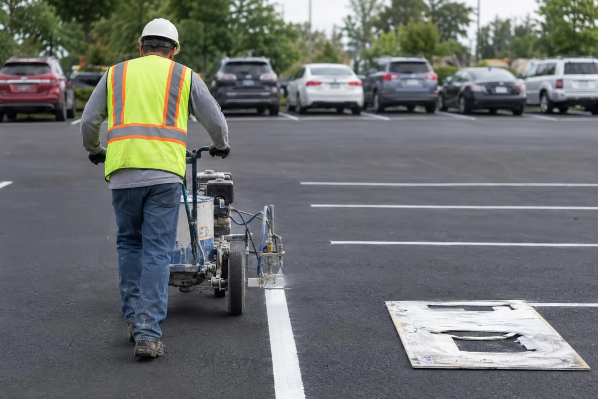Worker in a safety vest pushing a line striping machine to paint a white parking stall line